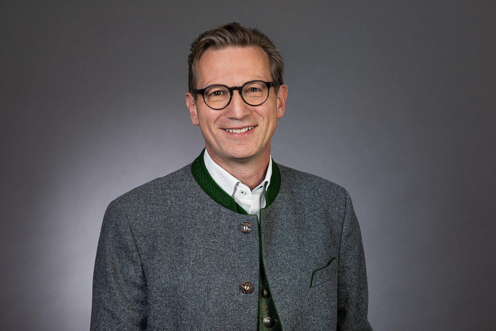 Alois Hager. Lachender Mensch in Tracht vor einer grauen Wand.